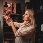 Beautiful focused woman is choosing right wine for customers at dark wine cellar.